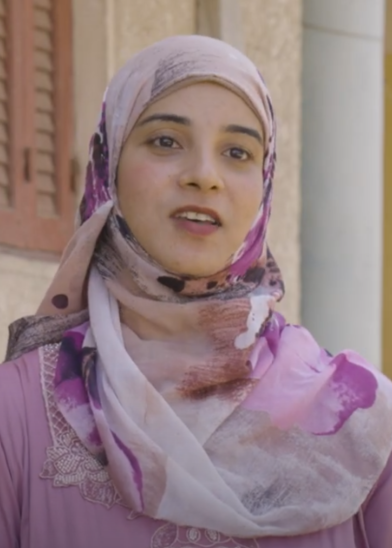 A young woman wearing a light pink and purple patterned hijab and a matching top with lace detail stands outside, speaking. She has light skin and dark eyes. Behind her is a beige wall, a window with wooden shutters, and a white column.