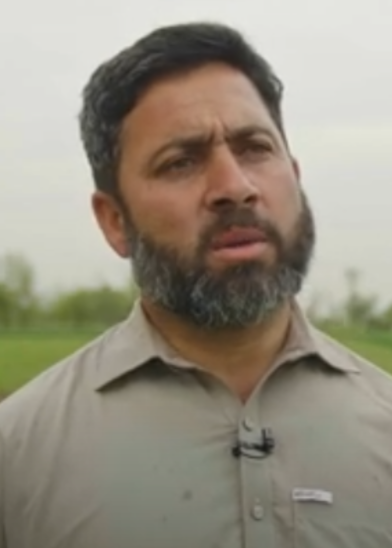 A man with short dark hair and a full beard stands outdoors in a field, wearing a light gray collared shirt. He has a serious expression on his face and a small microphone clipped to his shirt. The background is blurred with trees and a cloudy sky.