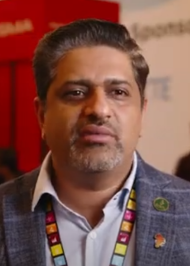A man with short, combed-back salt-and-pepper hair and a trimmed beard wears a checked blazer, white shirt, and colorful lanyard. He stands indoors in front of blurred expo signage and a red display, looking slightly past the camera.