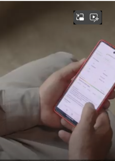 A person wearing light-colored clothing holds a red-bordered smartphone in both hands, browsing a document or web page with text and some charts or tables. The background is blurred, and part of a video player icon is visible on the screen.