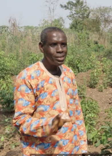 A middle-aged man stands outdoors in a lush, green field. He wears a colorful, patterned long-sleeve shirt with blue and orange designs. He gestures with one hand while speaking, and trees and plants are visible in the background under a bright sky.