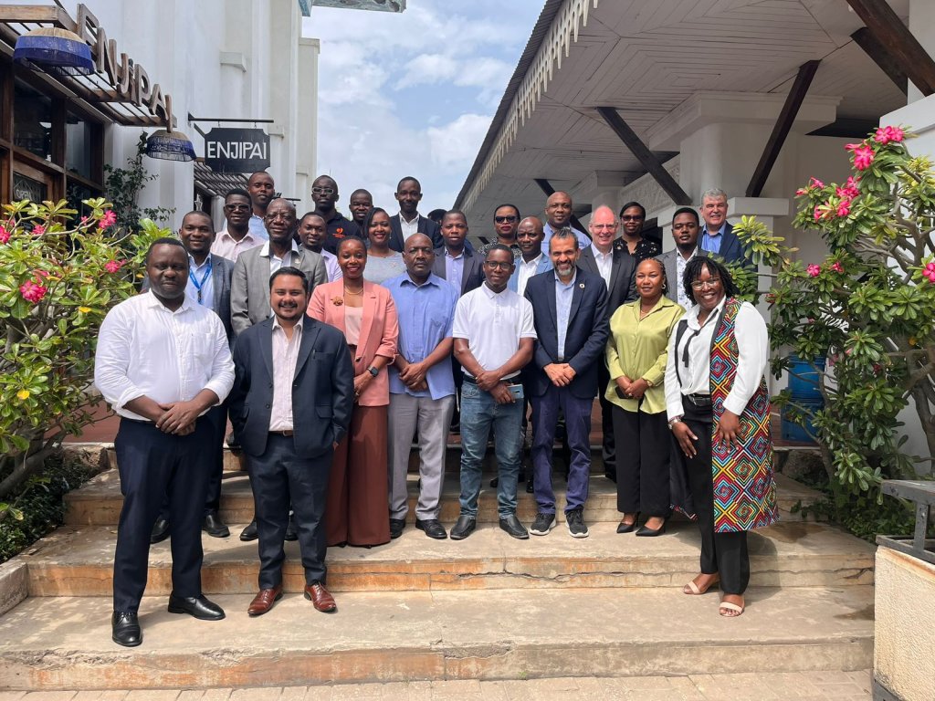 A group of about 25 people, both men and women, stand smiling on stone steps outside a white building with flowers and greenery. Wearing business attire, they gather for a photo after discussing life-saving early warning systems on a sunny day.