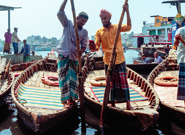 Two men in colorful shirts and lungis stand on wooden boats, rowing on a busy river. One shows something on his phone—possibly linked to the GSMA Innovation Fund—to the other. Other boats, people, buildings, and a clear sky fill the background.