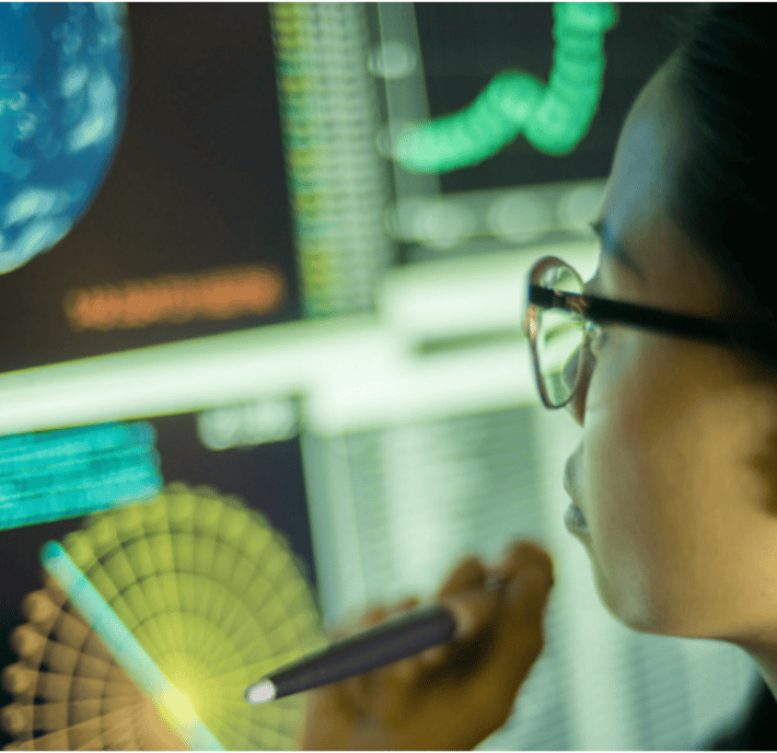 A person wearing glasses closely examines data and charts on multiple computer screens, holding a pen. The colorful graphs, digital globe, and complex visualizations suggest climate finance analysis or scientific research.