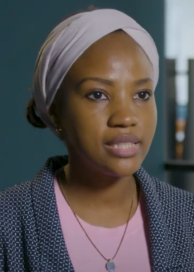 A woman wearing a pale headscarf, light pink top, and patterned blazer looks slightly past the camera. She has a short necklace with a round pendant. The background is out of focus, with blue walls and bookshelves visible.