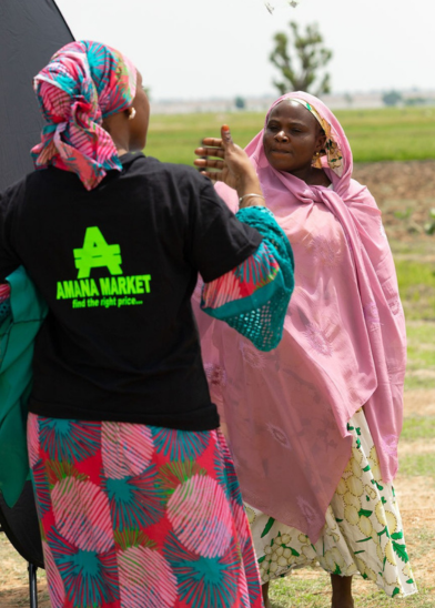 Two women stand outdoors on grassy land. One, wearing a black "AMANA MARKET" shirt, headscarf, and patterned skirt, faces away. The other, in a pink hijab and cream skirt, gestures with her hand, looking intently at the first woman. Trees and fields appear in the background.