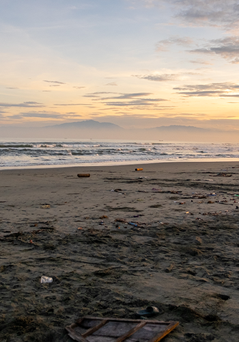 A sandy beach at sunset with scattered trash and debris along the shore. Waves gently roll in, and distant mountains are faintly visible under a cloudy sky with orange and blue hues. The scene highlights pollution on an otherwise scenic coastline.