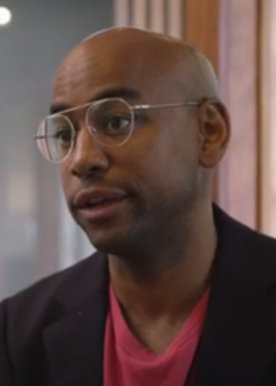 A man with a shaved head, wearing round glasses and a dark blazer over a bright pink shirt, is shown indoors. He appears to be speaking, with a neutral expression, and a softly lit background with wooden elements is visible.