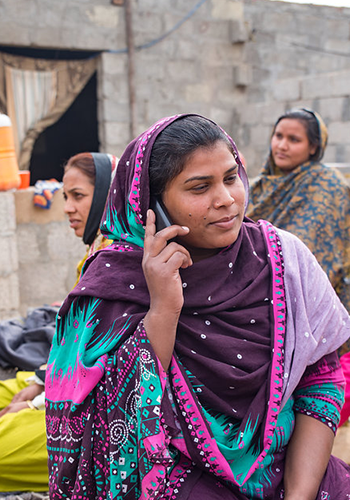 A woman in a colorful purple and teal patterned shawl talks on a mobile phone, sitting outdoors. Two women in patterned clothing and scarves sit behind her, with stone walls and a curtained doorway in the background. All appear to be engaged and relaxed.
