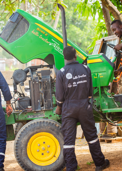 Three men wearing dark coveralls with “hello tractor” written on the back inspect and repair a green and yellow John Deere tractor. The engine cover is open. One man stands on the tractor, while two others examine its side under leafy trees outdoors.
