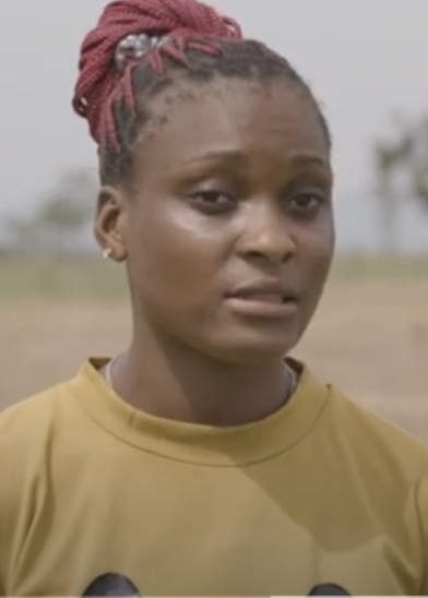 A young woman with braided red hair pulled up, wearing small earrings and a mustard yellow top, stands outdoors. She looks slightly to the side, with a neutral expression. The background is blurred, showing hints of trees and open land.