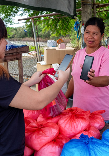 A woman in a pink shirt stands behind a table of red and blue plastic bags, smiling as she holds up her phone for another person, who is scanning it with their own phone. Cardboard boxes and a wire fence are visible in the background.