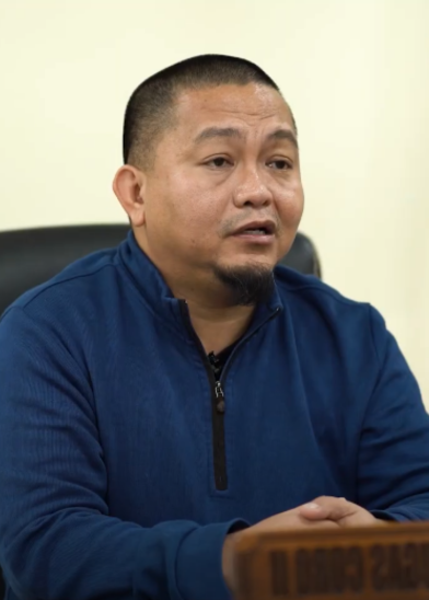 A man with short hair and a trimmed beard, wearing a blue zip-up sweater, sits at a desk with his hands clasped. He appears to be speaking or listening, and the background is neutral and out of focus.