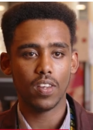 A young man with short, neatly shaped hair and a faint mustache looks slightly past the camera. He wears a collared shirt and jacket. The background is softly blurred, suggesting an indoor setting with warm lighting.