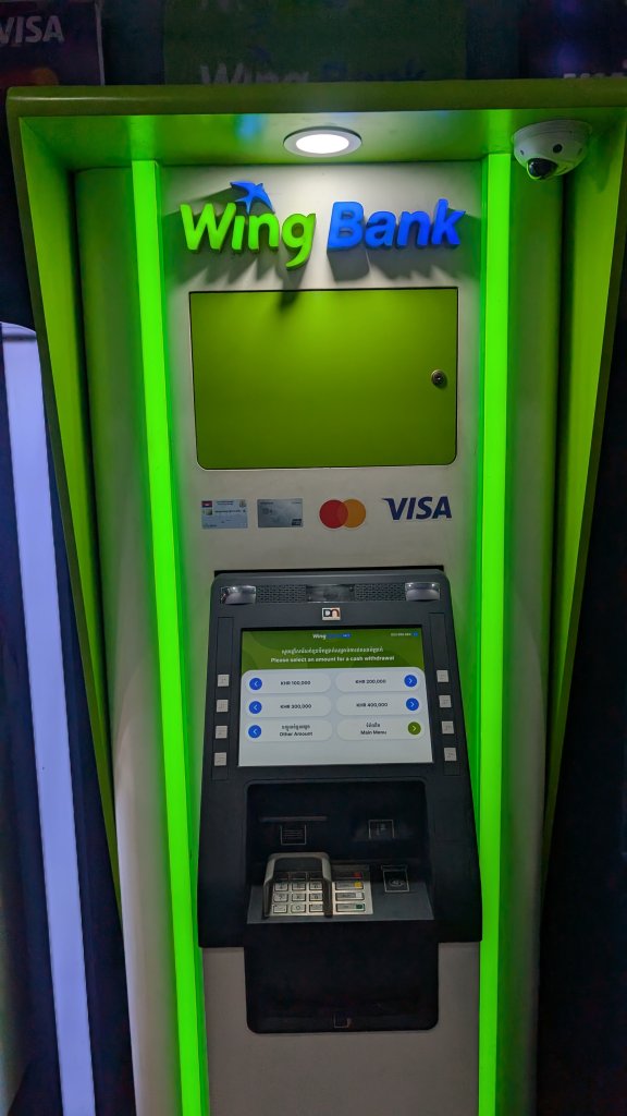 A Wing Bank ATM at airport is shown with a bright green frame. The machine features a screen, card slot, keypad, and cash dispenser. Logos for Mastercard and Visa are visible, and there is a security camera in the upper right corner. The ATM interface displays transaction options.