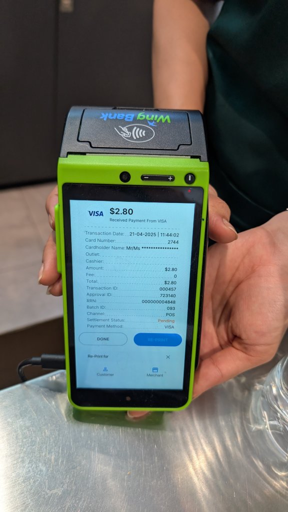 A person holds a green Wing Bank payment terminal at airport displaying a successful $2.80 VISA card transaction. The screen shows transaction details including date, card number, customer name, and a "Received Payment" confirmation.