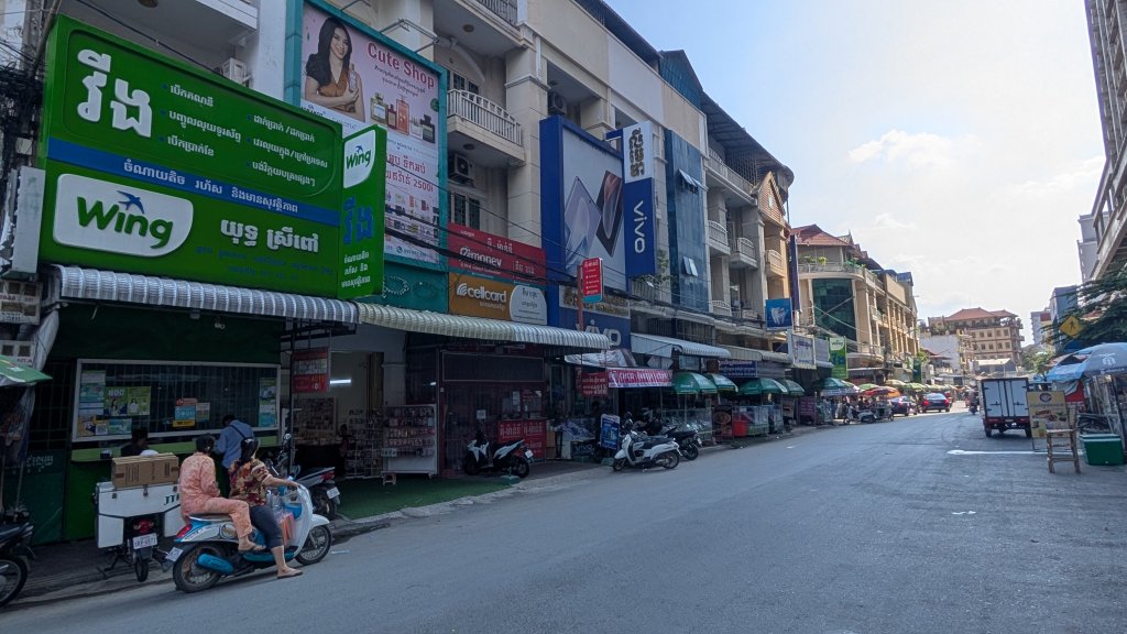 A busy urban street lined with multi-story shops, bright signs in Khmer and English, parked motorbikes, and people. Two women ride a scooter in the foreground. The sky is mostly clear, and sunlight casts strong shadows on the buildings and road.