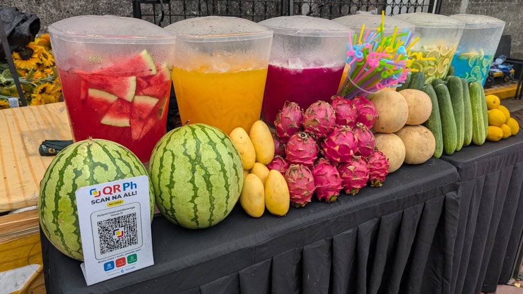 Colorful juice containers with vibrant drinks sit behind fresh fruits—watermelon, mangoes, dragon fruit, melons, and cucumbers. A QR code sign for GCash promotes financial inclusion alongside colorful straws on the table.