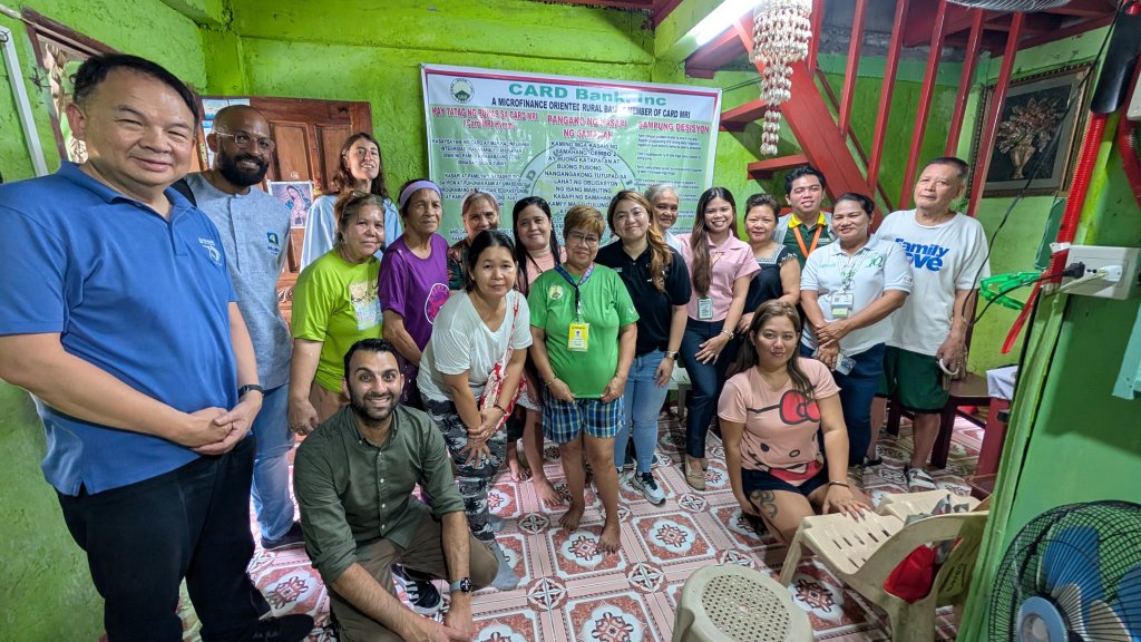 A diverse group of people pose and smile inside a small, green-walled room with patterned floor tiles. A large banner for CARD Bank, known for promoting financial inclusion, hangs at the back, indicating a community or outreach event.