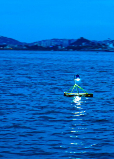 A small floating device with neon yellow supports and a blue flag emits light on a calm, blue-toned body of water at dusk. City buildings and hills are visible in the distant background under a deep blue sky.