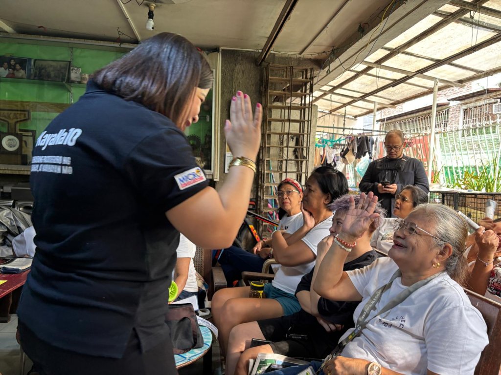 A woman in a black shirt gives a high-five to an older woman in glasses who is seated and smiling. Other elderly women, also seated, watch and smile. The group is under a covered outdoor area with various household items in the background.