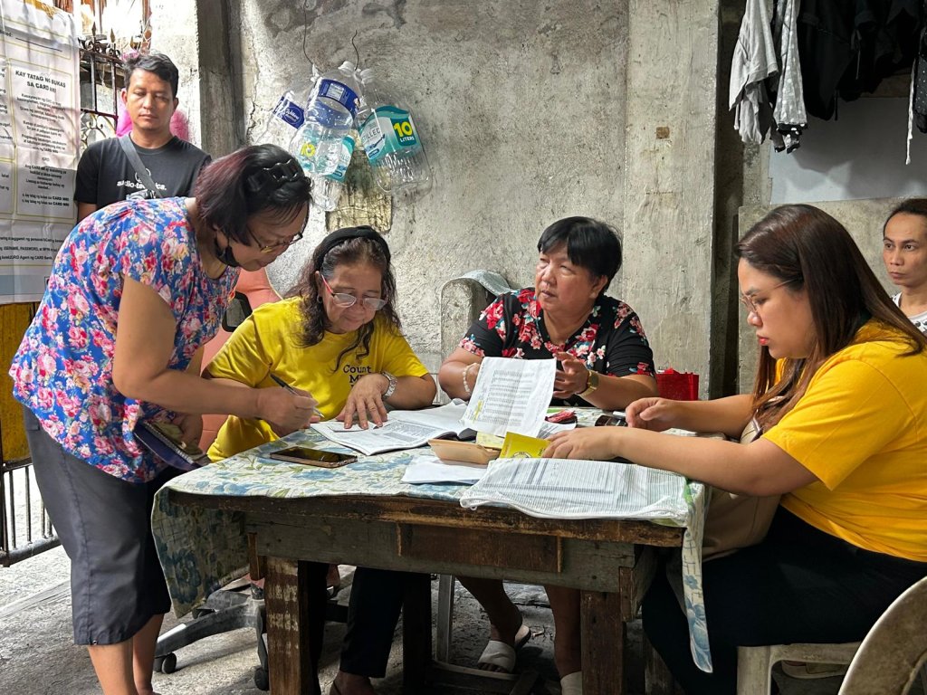 Four women sit around a worn table covered with papers, forms, and pens in a rustic indoor setting. One woman stands and signs a document while others assist her. A man and two women stand in the background. The mood appears focused and collaborative.