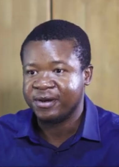A man with short black hair and medium-dark skin wears a royal blue button-up shirt. He looks slightly to the side with a neutral expression. The background is softly blurred and beige, possibly an indoor setting with wooden panels.