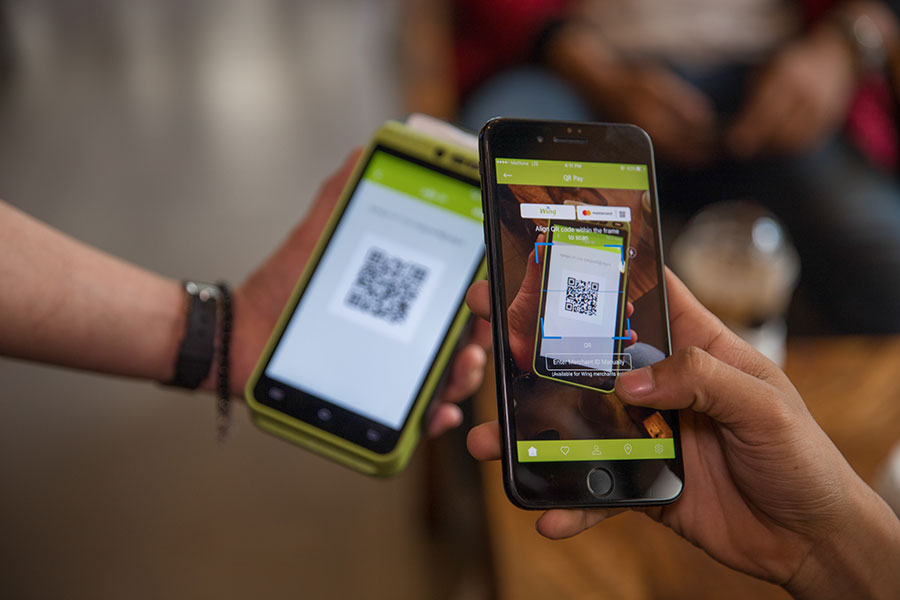 Close-up of two hands holding smartphones. One phone displays a QR code, while the other phone is using its camera to scan the code. The background is blurred, suggesting a casual indoor setting, possibly a café or shop.