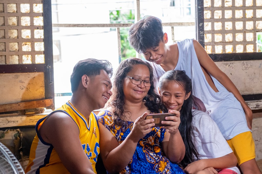 A woman sits smiling with three teenagers around her, all happily looking at a smartphone she is holding. They are indoors by a window with sunlight coming in. The group appears cheerful and close, sharing a joyful moment together.