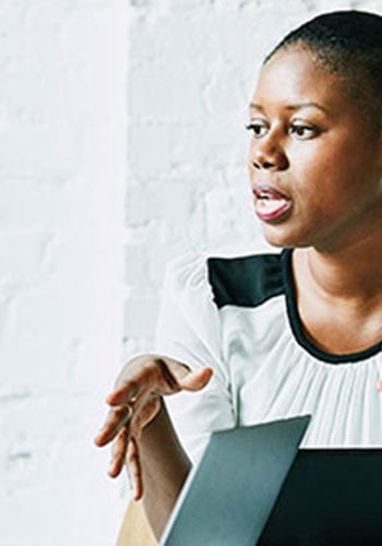 A woman with short hair wearing a white blouse with black accents gestures with her hands while speaking. She appears engaged in conversation, sitting indoors near a white brick wall and in front of a laptop.