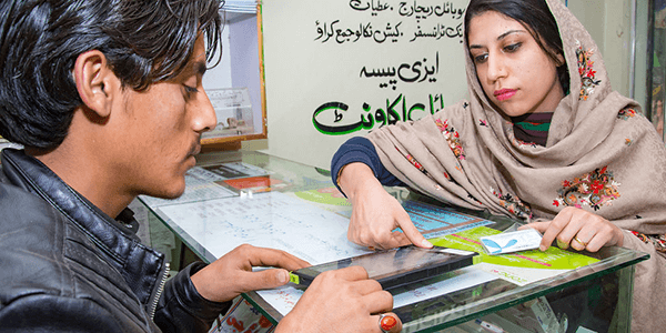 A woman in a light, patterned headscarf points to a digital tablet on a glass counter while a man interacts with the screen. Papers and informational brochures are on the counter, with text in Urdu on the background wall.