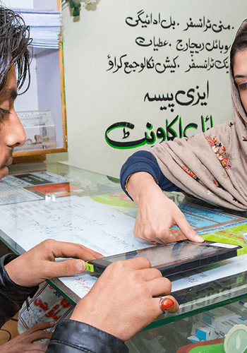A woman in a patterned headscarf points at a tablet screen on a counter, showing something to a man in a leather jacket. Behind them is a wall with Urdu text and brochures, suggesting a service or information desk setting.