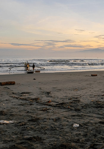 A sandy beach at sunset with scattered debris in the foreground. Two people stand near a small fishing boat by the shoreline, facing gentle waves. The sky is orange and blue with wispy clouds, and distant mountains are visible on the horizon.