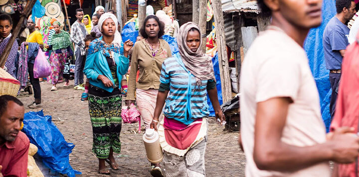 A bustling outdoor market scene with many people walking, wearing colorful clothing. Some women carry items on their heads or hold containers. The street is crowded and lively with stalls and tarps lining the way.
