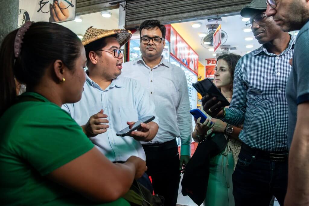 A group of six adults stands together in a brightly lit shop or market area. One man in a straw hat and glasses gestures while holding a phone; others listen or look at their own devices. The mood is engaged and conversational.