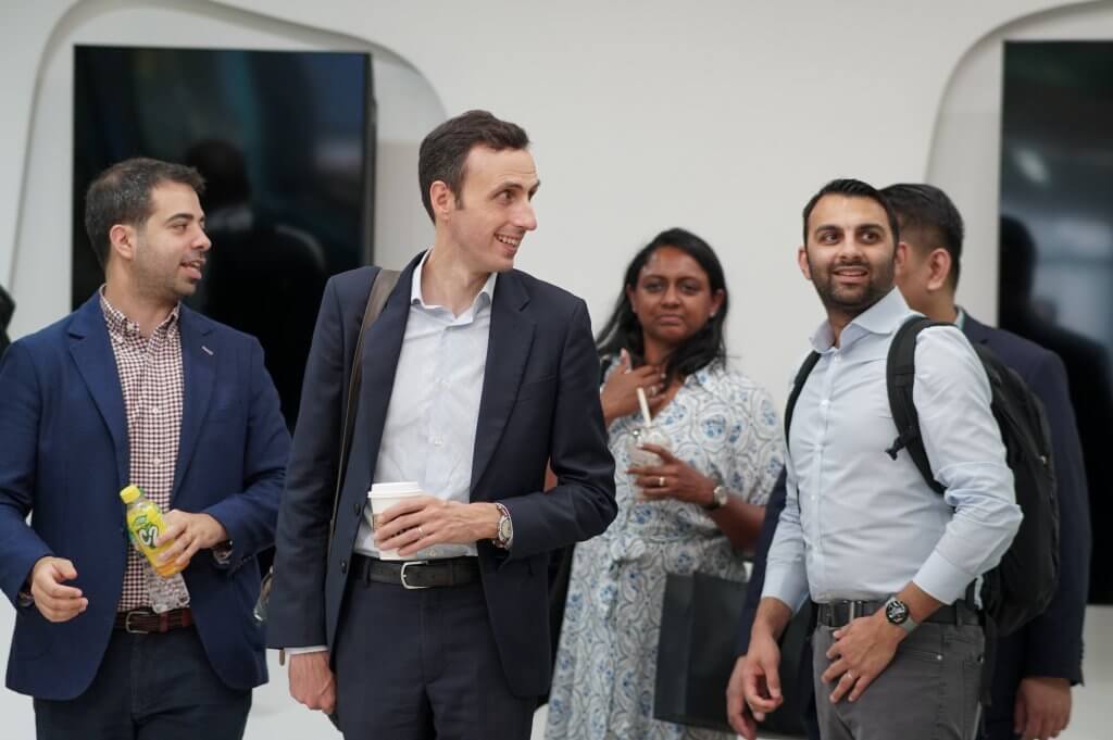 Five professionally dressed people stand indoors, smiling and conversing. One holds a beverage bottle, another a coffee cup and backpack. A woman in a patterned dress is in the background. The setting is modern, with white walls and large TV screens.