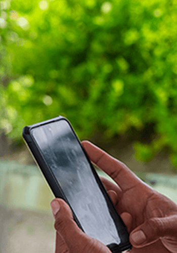 A close-up of two hands holding and using a smartphone with a black case. The background is blurred, showing bright green foliage and suggesting an outdoor setting. The phone's screen is slightly reflective.