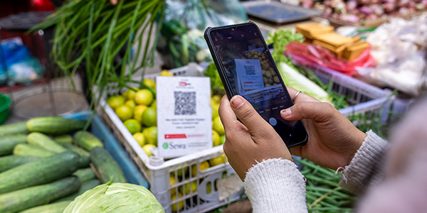 A person holds a smartphone and scans a QR code displayed on a sign at a vegetable market stall, surrounded by fresh produce like lemons, cucumbers, and cabbage. The QR code likely enables digital payment for the produce.