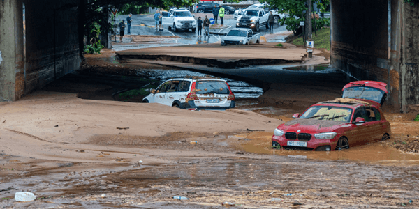 Flooded underpass with several partially submerged cars, mud covering the ground, and damaged road. One red car has its trunk open; people and vehicles are gathered on drier ground in the background, observing the scene and damaged area.