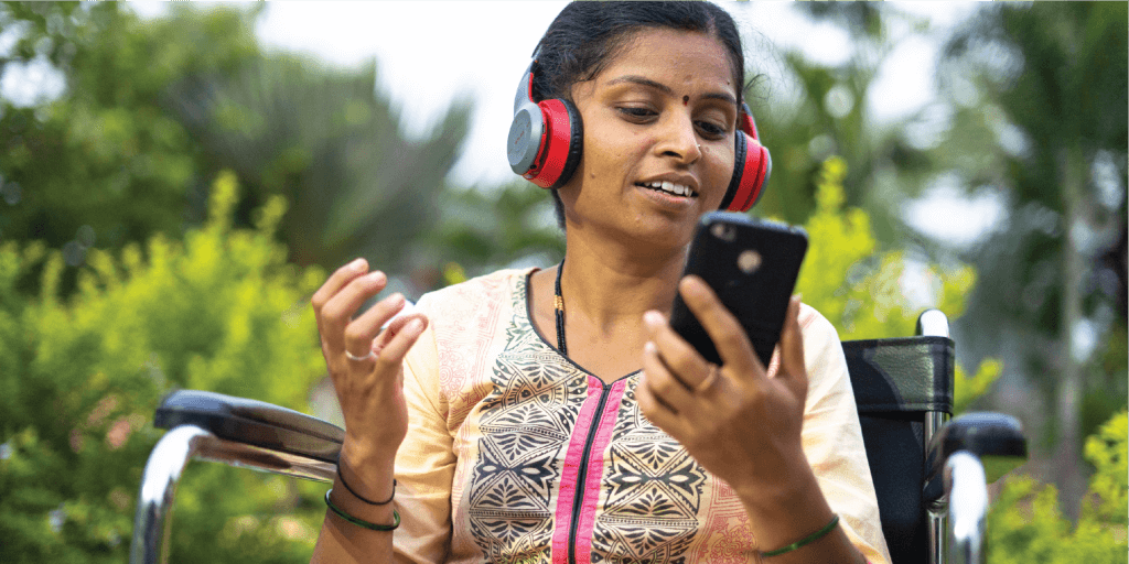 A woman in a wheelchair sits outdoors, bridging the mobile disability gap as she smiles with red and black headphones on, holding a smartphone and gesturing happily. She wears a patterned peach and pink kurta, surrounded by green plants and trees.
