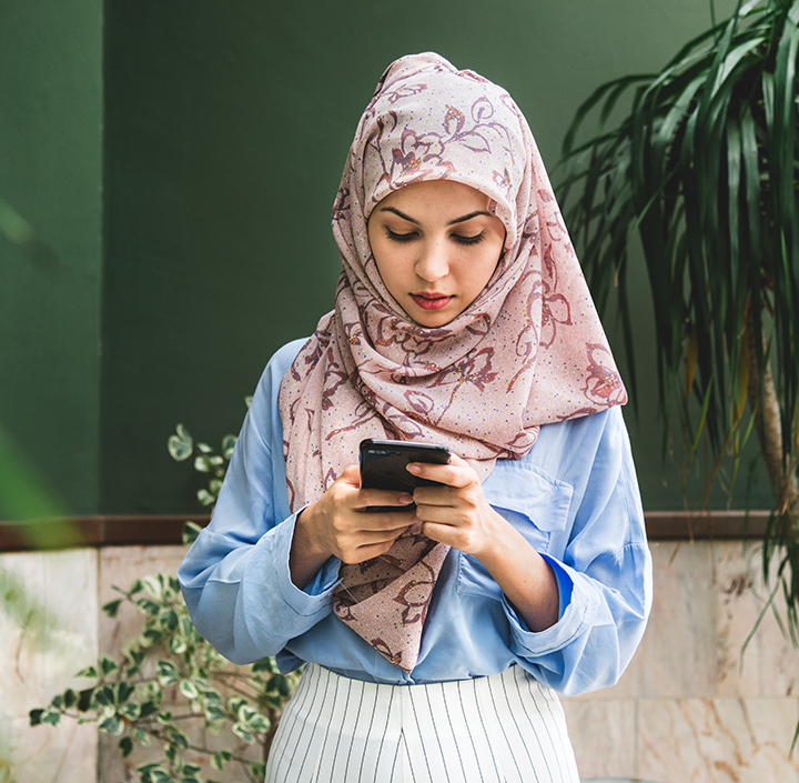 A young woman wearing a light pink patterned hijab and a blue shirt stands indoors, looking down at her phone. Near green plants by a green wall, she appears focused on her device, reflecting the impact of GSMA Mobile for Development initiatives.