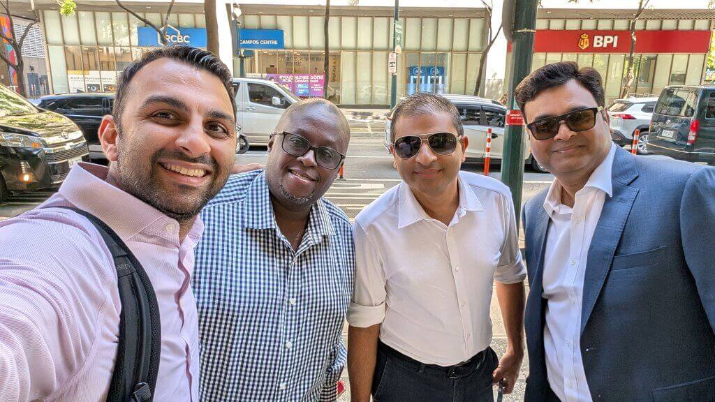 Four men wearing collared shirts and glasses, some in business attire, smile for a selfie on a city sidewalk. Behind them are parked cars and buildings, including banks with blue and red signs reading “RCBC” and “BPI.” It’s a sunny day.