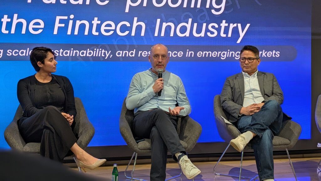Three people sit on stage in modern chairs, participating in a panel discussion. The man in the center holds a microphone and speaks, while the woman on the left and the man on the right listen. A large blue screen behind them displays fintech industry-related text.
