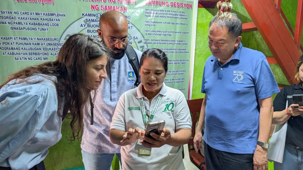 Four adults stand together indoors. A woman in a white polo shirt shows something on her phone to two people leaning in closely; a woman with long brown hair and a man in glasses. Another man in a blue shirt stands nearby. Large posters in Filipino hang on the green wall behind them.