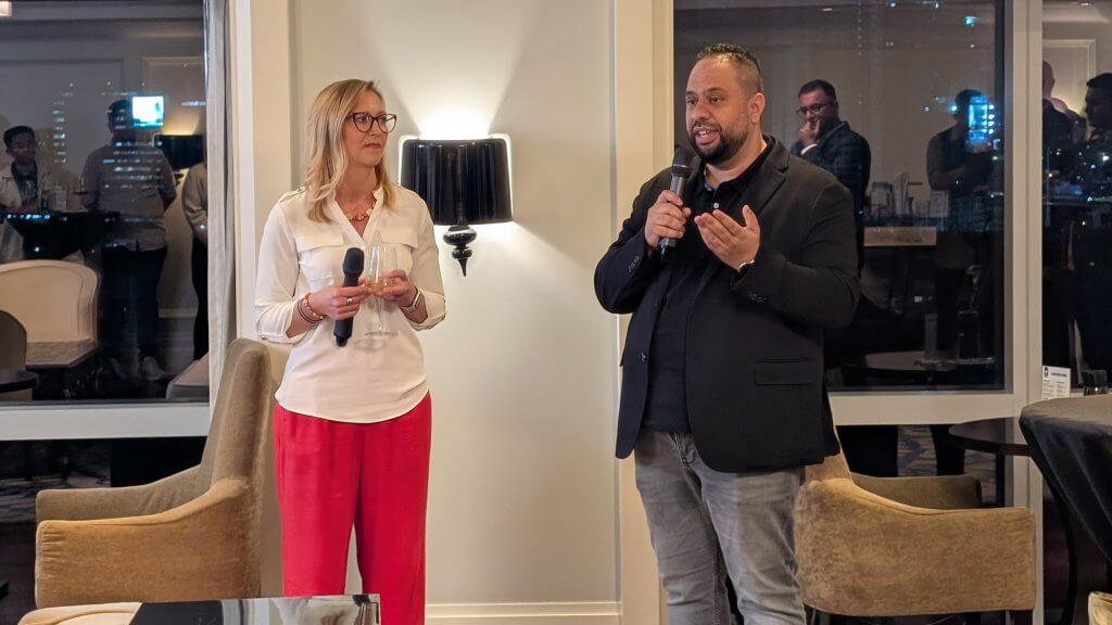 A woman in a white blouse and red pants stands holding a microphone, looking at a man in a black jacket and jeans who speaks into a microphone. They are indoors, with seated guests and city lights visible through large windows behind them.
