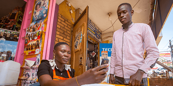Two people are at a small shop. One, seated and wearing an orange apron, looks at a smartphone, while the other, standing in a pink shirt, watches. The shop has hair product posters and colorful items on display.