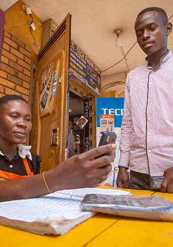 A person sitting at a yellow table uses a mobile phone while another person stands beside them. They are indoors, with a logbook, documents, and tech advertisements visible in the background. Both appear focused on the phone.