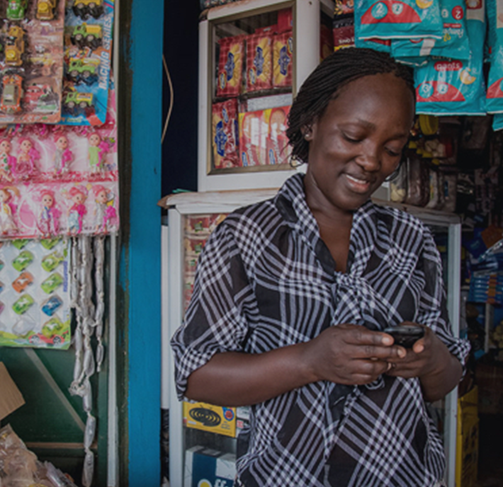 A woman wearing a black and white plaid shirt stands smiling while using a smartphone. She is in front of a colorful shop display with various packaged goods, including snacks and toys, hanging and stacked behind her.