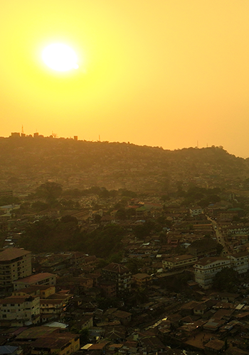 A golden sun sets over a densely populated hillside city. The sunlight casts a warm yellow-orange glow across the sky and buildings, creating long shadows. Clusters of houses and winding streets are visible in the urban landscape below.