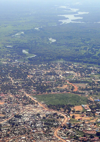 Aerial view of a town with clustered buildings, dirt roads, and patches of greenery. A river flows through lush, green landscape in the background, winding towards the horizon under a hazy sky. The area appears rural with scattered settlements and open fields.